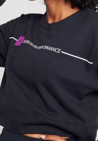 Zwart cropped sweatshirt met de tekst "Calvin Klein Performance" en een paarse vierkante accent, gedragen door een persoon met de hand op de heup.