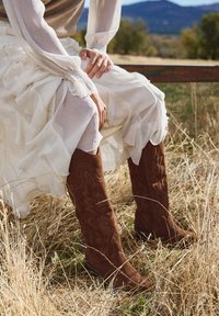 Braune Wildlederstiefel mit bestickten Akzenten, hohem Schaft und flacher Sohle, kombiniert mit einem mehrlagigen weißen, gerafften Rock und einem durchsichtigen Oberteil.