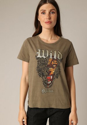 Femme portant un t-shirt vert olive avec un graphique moitié léopard, moitié tigre et le texte "Esprit Sauvage" à l'avant, se tenant devant un fond neutre.