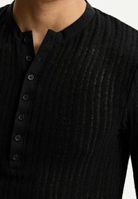 Camisa henley negra con textura acanalada, un cuello redondo y una tapeta con seis botones negros. El material parece ligero y ligeramente transparente.