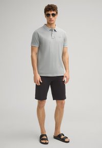 Lichtgrijze poloshirt met een kraag, gecombineerd met zwarte shorts. Het model draagt donkere zonnebril en zwarte sandalen voor een casual look.