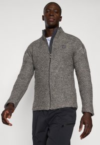 Chaqueta gris con cremallera y un tejido texturizado, que cuenta con un cuello alto y un parche con el logo en el pecho. Acentuada con costuras tonales en las mangas.