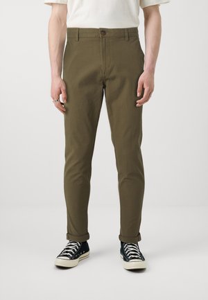 Homme portant un pantalon slim vert olive retroussé aux chevilles, une chemise blanche à manches courtes, des baskets montantes noires et des bagues aux doigts.