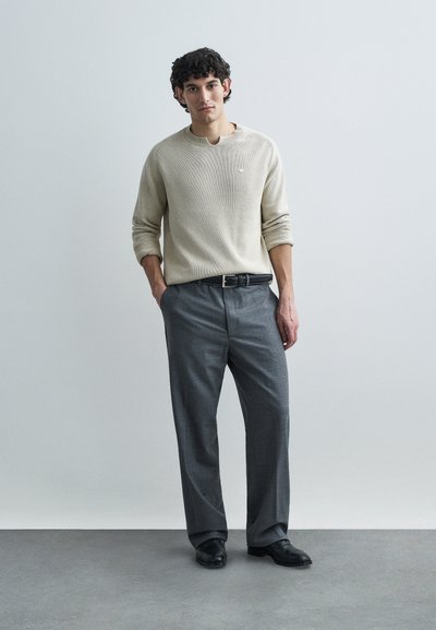 Μπεζ ριμπ πλεκτό πουλόβερ με στρογγυλή λαιμόκοψη, σε συνδυασμό με γκρι πλισέ παντελόνια και μαύρα παπούτσια, παρουσιάζονται σε ουδέτερο φόντο.