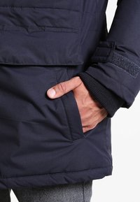 Marinblå isolerad jacka med dragkedja, sidofickor och fleecefoder på ärmsluten; har ett texturerat tyg och en kardborreknapp på ärmen.