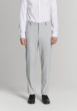 Homme portant un pantalon gris clair sur mesure, une chemise blanche et des mocassins en cuir noir, debout devant un arrière-plan gris clair uni.