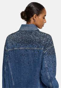 Giubbotto in denim con un effetto blu lavato, impreziosito da strass neri e argento sulle spalle e sulla parte superiore della schiena, colletto classico e chiusura con bottoni.