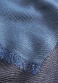 Écharpe en tissu bleu tissée avec des extrémités à franges, ornée d'un motif diagonal. La texture semble douce, superposée à un tissu texturé plus foncé.