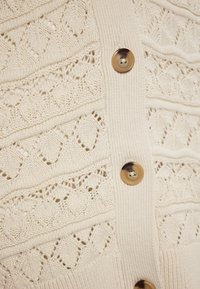 Gebreide cardigan in crème met ingewikkelde opengewerkte patronen, voorzien van goudkleurige knopen en ribbels aan de randen voor textuur en detail.