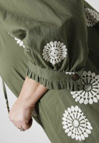 Olijfgroene blouse met witte geborduurde bloemenpatronen, ruches aan de mouwen en een knoopdetail. Zachte stof met een losvallend ontwerp.