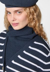 Marine gebreide trui met een hoge, geribbelde kraag, voorzien van witte horizontale strepen. Zachte textuur met een asymmetrisch ontwerp en details in goudkleurig hardware.