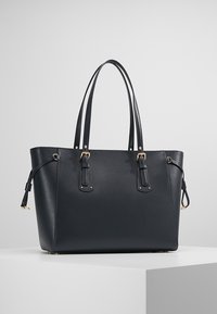 Suuri musta nahkainen olkalaukku, jossa on kultaiset yksityiskohdat, jämäkkä muoto, kaksi kahvaa ja koristeelliset yksityiskohdat sivuilla.