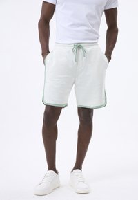 Pantaloni corti in cotone grigio chiaro misto con bordi verdi e coulisse, dotati di tasche laterali e taglio classico. Indossati con sneakers bianche.