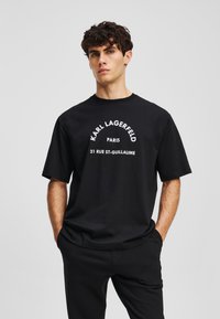 Juodas medvilninis marškinėlis su balta užrašu "KARL LAGERFELD PARIS 21 RUE ST-GUILLAUME", išspausdintu kreivais, drąsiais šriftu. Trumpomis rankovėmis ir apvalia v-neck.