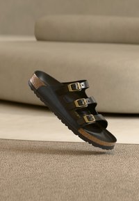 Sandale noire avec trois boucles dorées réglables et semelle en liège flottant au-dessus d'un tapis beige dans une pièce aux tons neutres.