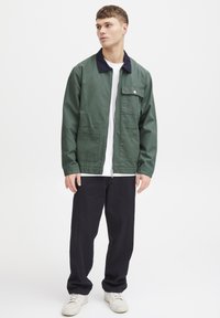 Grøn letvægtsjakke med en marineblå krave, frontlynlås og brystlomme. Båret over en hvid t-shirt og parret med mørke bukser. Hvide sneakers.