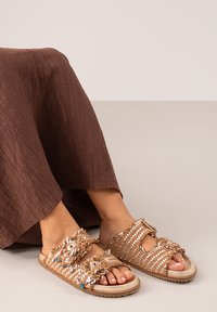 Sandalias deslizantes de color marrón, tejidas, que presentan embellecedores decorativos, correas texturizadas y una suela flexible para mayor comodidad.