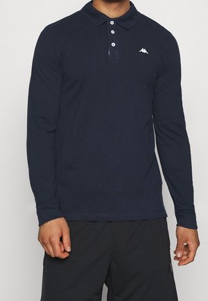 Mann trägt ein marineblaues Langarm-Poloshirt mit drei weißen Knöpfen und kleinem weißem Logo auf der linken Brust, kombiniert mit schwarzer Hose.