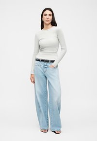 Femme portant un haut à manches longues gris clair, un jean large bleu clair, une ceinture noire et des talons ouverts noirs, debout avec une main dans la poche.