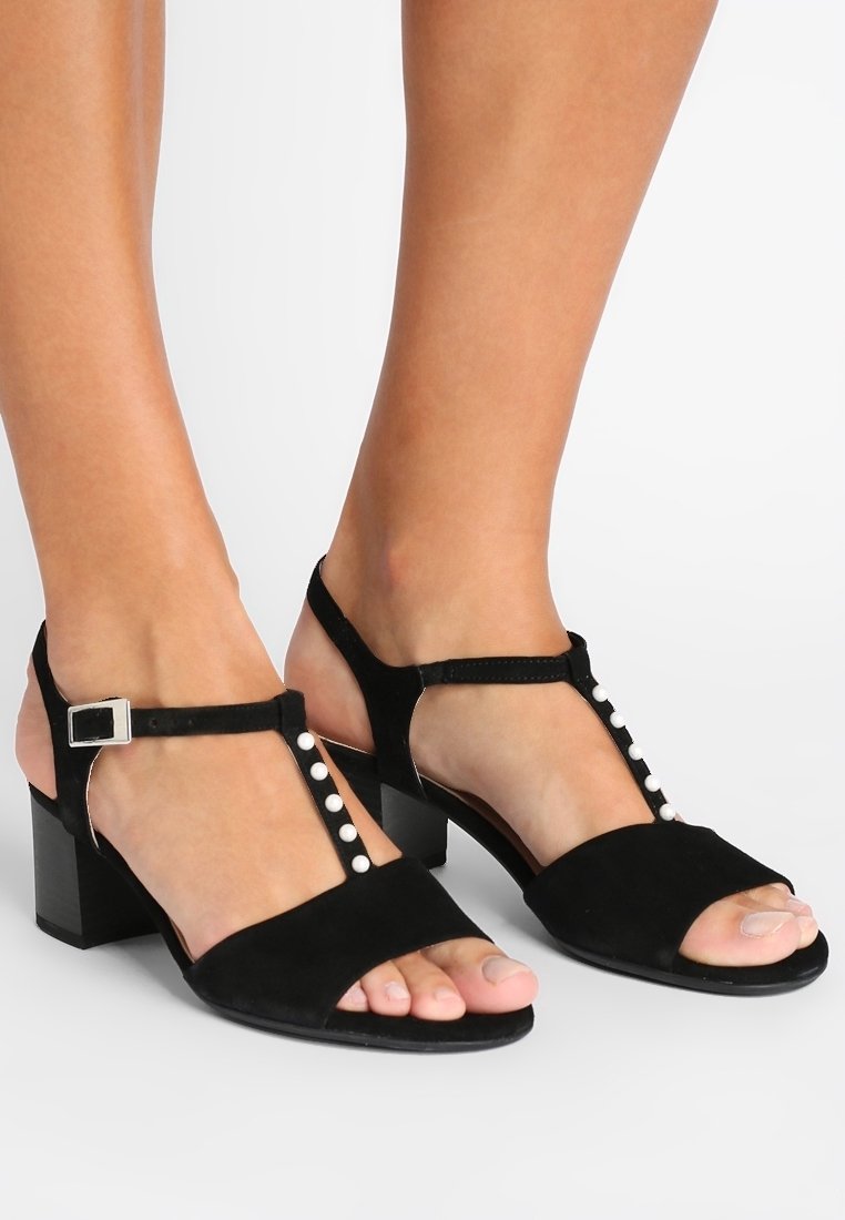 Pieds portant des sandales à brides en T en daim noir avec des talons blocs bas et de petits détails de boutons blancs sur les brides.