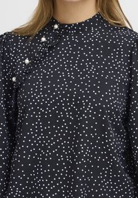 Blusa nera con pois bianchi, caratterizzata da un lato sinistro arricciato e bottoni simili a perle al collo. Tessuto leggero con una textura liscia.