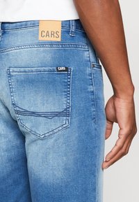 Denim jeans in light blue wash, featuring a back pocket with a black tag reading "CARS" and a tan leather back patch with "CARS."