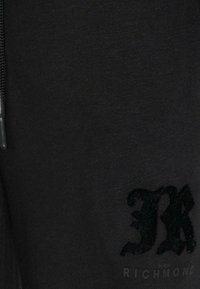 Czarna bluza z teksturowanym nadrukiem litery „K” oraz nazwą marki „Richmond” w subtelnym, ciemnym wzorze. Miękki materiał, swobodny krój.