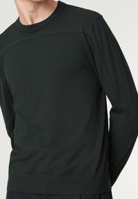 Chemise à manches longues vert foncé en tissu doux, avec un col rond et des détails de couture contrastants le long des épaules.