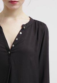 Blusa nera con bottoni a pressione, scollo a V e maniche corte plissettate. Realizzata in un tessuto leggero e morbido.