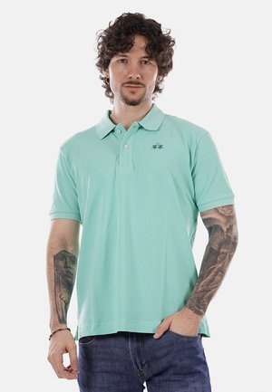 Uomo con capelli ricci che indossa una polo verde chiaro e jeans, con tatuaggi dettagliati su entrambi gli avambracci, in piedi con una mano in tasca.