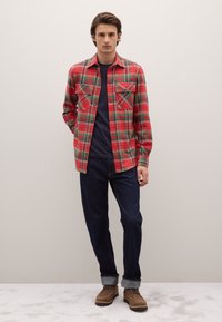 Camicia a quadri rossa con bottoni e due tasche sul petto, abbinata a una t-shirt blu navy con scollo tondo, jeans blu scuro e stivali marroni con lacci.