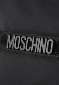 Sort nylon taske med en flad strop og et hævet, mat sort "MOSCHINO" logo. Glat tekstur med subtilt væve mønster.