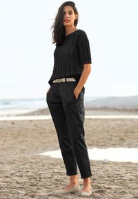 Schwarzes gestricktes Kurzarmoberteil, beiger Gürtel, lockere schwarze Hose und hellbeige Ballerinas. Sandstrand im Hintergrund, sanfte Wellen sichtbar.