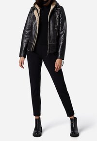 Giacca nera in pelle con fodera in pelliccia sintetica, chiusura a zip e cappuccio. Abbinata a pantaloni neri su misura e stivaletti neri alla caviglia.