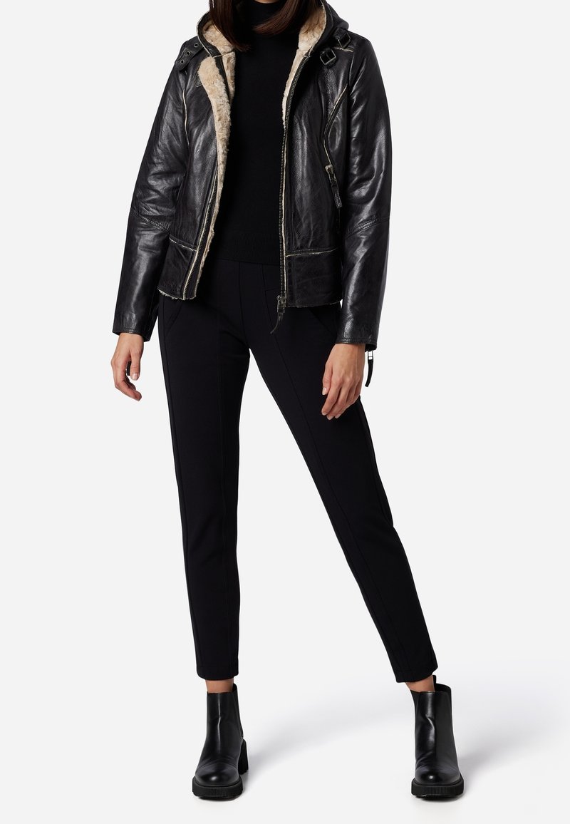 Giacca nera in pelle con fodera in pelliccia sintetica, chiusura a zip e cappuccio. Abbinata a pantaloni neri su misura e stivaletti neri alla caviglia.