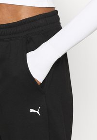 Zwarte joggers met een elastische tailleband, voorzien van een zijzak en een wit Puma-logo. Een wit langetermijn top is gedeeltelijk zichtbaar.