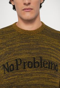 Tekstuirani pleteni pulover u smeđoj i senf žutoj boji središnjim crnim natpisom "No Problemo." Okrugli izrez.