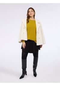 Cappotto bianco doppiopetto, maglione giallo lavorato a maglia, gonna nera a pieghe, calze nere e stivali neri al ginocchio con finitura lucida.