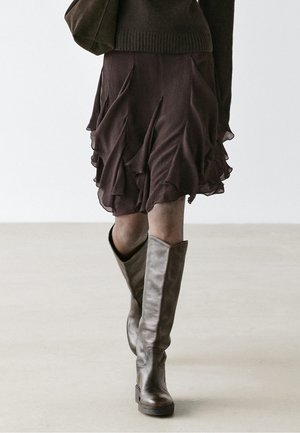 Person trägt einen dunklen, rüschigen Rock und braune kniehohe Lederstiefel und läuft auf einem schlichten hellen Boden vor einem weißen Hintergrund.