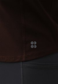Tamno smeđa sportska majica s glatkom teksturom i ravnim šavovima, sa malim sivim logotipom na donjoj strani. Crne tajice s kontrastnim šavovima.