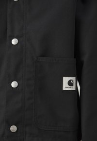 Veste noire avec des boutons-pression en métal, une poche poitrine et un logo Carhartt bien visible dans un carré contrasté. Texture de tissu durable.