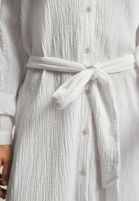 Chemise blanche texturée avec un devant à boutons et des manches longues, cintrée à la taille avec une ceinture assortie.