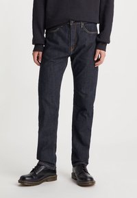 Person som bär mörkblå raka jeans, svarta läder snörskor och en svart långärmad topp, ståendes mot en vit bakgrund.