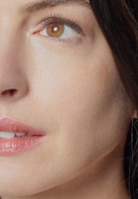 Nærbilde av et ansikt som viser glatt, lys hud, en lett glans på leppene, og et brunt øye med en sirkulær refleksjon. Subtil hårtekstur er synlig.