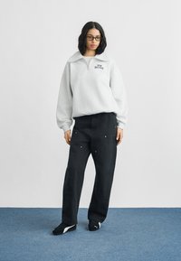 Γυναίκα με γκρι φούτερ New Balance με φερμουάρ μέχρι τη μέση, μαύρο παντελόνι φαρδιάς γραμμής, μαύρα αθλητικά παπούτσια Puma και γυαλιά, στέκεται σε μπλε χαλί.