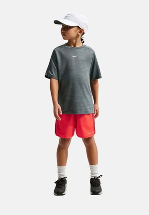 Niño con gorra blanca, camiseta gris de Nike, pantalones cortos rojos, calcetines blancos y zapatillas negras, de pie y mirando hacia un lado.