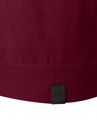 Närbild av en ribbad fåll på ett vinrött stickat plagg med en liten svart tygetikett sydd i nederkanten.