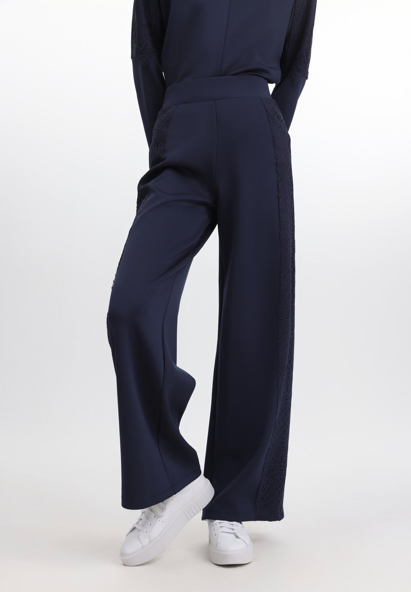 Pantaloni largi bleumarin, cu panouri din dantelă texturată pe laturi. Material moale, cu o finisare netedă. Asortați cu adidași albi.