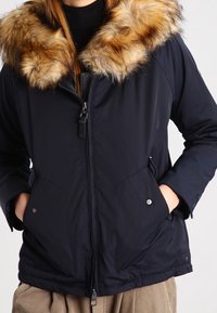 Laivansininen takki, jossa on pehmeä, vaaleanruskea turkiskalvo; varustettu vetoketjulla, kahdella sivutaskulla ja tekstuuripinnoitteella.