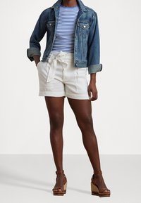 Džínová bunda, modro-bílý pruhovaný top, smetanové kraťasy s uvázaným páskem, hnědé sandály na klínu. Detaily zahrnují ohrnuté rukávy a texturovanou tkaninu.